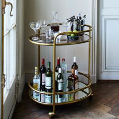 a gold bar cart with wine bottles and glasses on it in front of a window