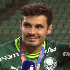 a man holding a microphone in his right hand and wearing a green shirt with silver medals around his neck