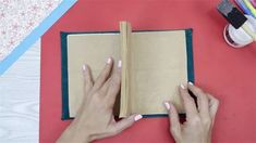 two hands are holding an open book on a table next to some markers and pencils