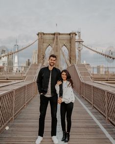 a man and woman standing on a bridge in front of the brooklyn bridge, looking at the camera