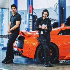 two people standing next to an orange sports car