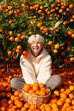 an older woman sitting on the ground surrounded by oranges in a basket and smiling