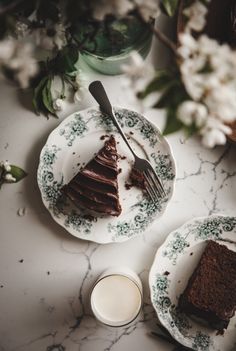 two plates with slices of chocolate cake on them next to a glass of milk and flowers