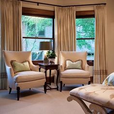 a bedroom with two chairs and a bed in front of three large windows that have curtains on them