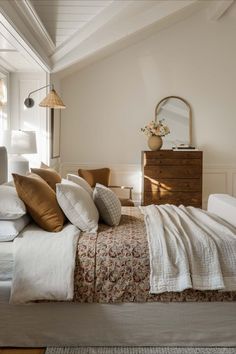 a bed with white sheets and pillows in a bedroom next to a dresser, mirror and lamp