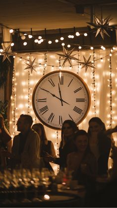 A large clock with Roman numerals on a wall adorned with string lights and star decorations at a festive gathering.