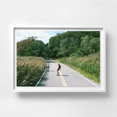 two skateboarders riding down the road in front of some tall grass and trees