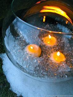 three lit candles are floating in ice and water inside a bowl on the ground with snow around it