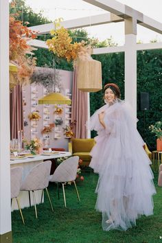 a woman wearing a white dress standing in the grass near a table with flowers on it