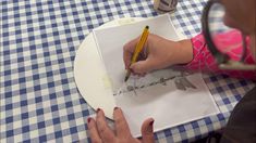 a woman is drawing on a piece of paper at a table with a blue and white checkered tablecloth