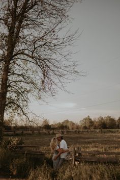 a man and woman sitting on a wooden bench in the grass near a tree with no leaves