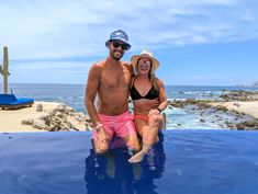 a man and woman sitting on the edge of a swimming pool in front of an ocean