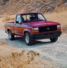 a red pick up truck is parked on the side of a dirt road near some dry grass