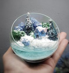 a hand holding a glass bowl filled with snow covered trees and houses in the snow