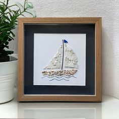 a white sailboat in a wooden frame on a table next to a potted plant