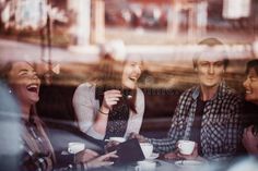 four people sitting at a table laughing and drinking coffee in front of a store window