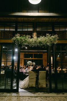 a couple sitting at a table in front of a building with flowers hanging from the ceiling