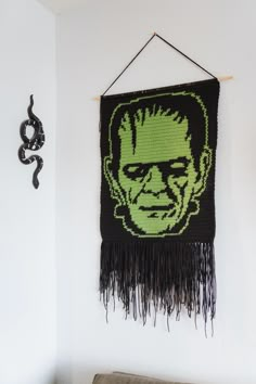 a crocheted wall hanging with a green face on it and black fringes