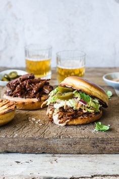 two pulled pork sandwiches with pickles on a wooden cutting board next to beer glasses