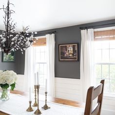 a dining room table with white flowers and candles on top of it, in front of two windows