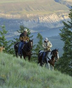 two men riding horses down a hill with mountains in the background