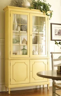 a yellow china cabinet with glass doors and plants on top in a dining room area