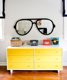 a yellow and white dresser with two mirrors on it's face, next to a pair of sunglasses