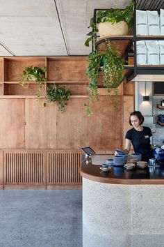 a woman standing behind a counter in a restaurant with plants hanging from the wall above it