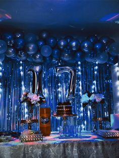 a table topped with lots of silver balloons and cake next to a wall covered in blue tinsel