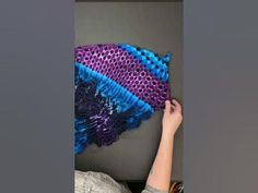 a woman is holding up a purple and blue fish netted wall hanging on a gray wall