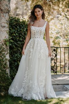 a woman wearing a wedding dress standing in front of a tree and bushes with her hand on her hip