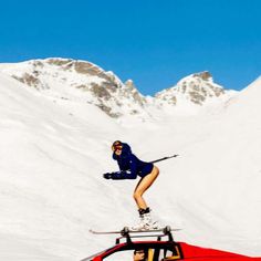 a woman on skis jumping over a car in the snow with mountains behind her