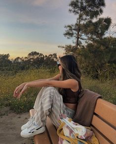 a woman sitting on top of a wooden bench next to a field with trees and grass