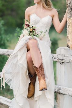 a woman in cowboy boots is sitting on a fence with her dress flowing over her