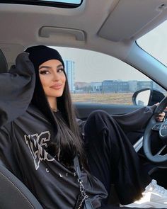 a woman sitting in the driver's seat of a car wearing a black hoodie