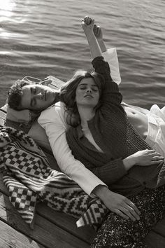 a man and woman laying on top of a wooden dock next to the water with their arms around each other