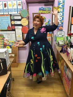 a woman in a blue dress is holding her hands up and standing in front of a classroom