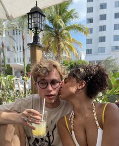 a man and woman kissing while holding a drink in front of their face with palm trees behind them