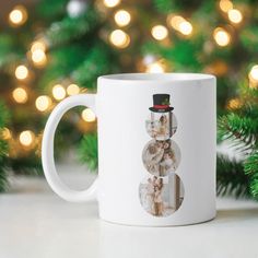 a white coffee mug sitting on top of a table next to a christmas tree with lights