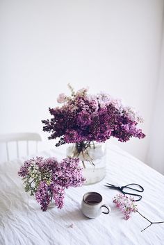 purple flowers in a vase and scissors on a white table cloth next to a cup of coffee