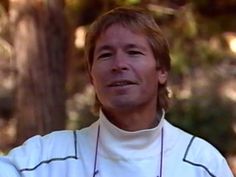 a man with long hair wearing a white shirt and black necklace standing in front of trees