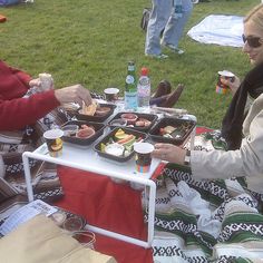 two women sitting at a picnic table with food and drinks in front of them on the grass