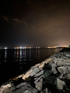 the night sky is lit up over the water and rocks on the shore with lights in the distance