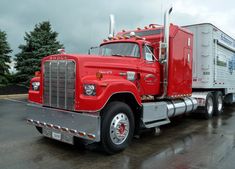 a red semi truck parked in a parking lot on a rainy day with trees behind it