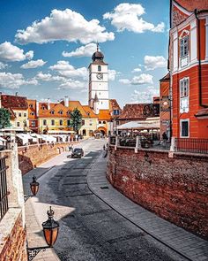 a clock tower towering over a city next to a brick wall and street light with cars driving on the road