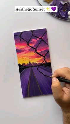 someone is painting a sunset on a piece of paper with purple and orange colors in the background