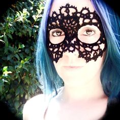 a woman with blue hair wearing a black lace mask on top of her face in front of bushes