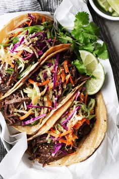 three tacos with shredded beef, cabbage and carrots on a white plate next to a bowl of cilantro