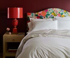 a red lamp is next to a bed with white sheets and floral headboard on it