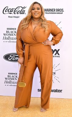 a woman in an orange jumpsuit and gold necklace standing on a yellow carpet with her hands on her hips